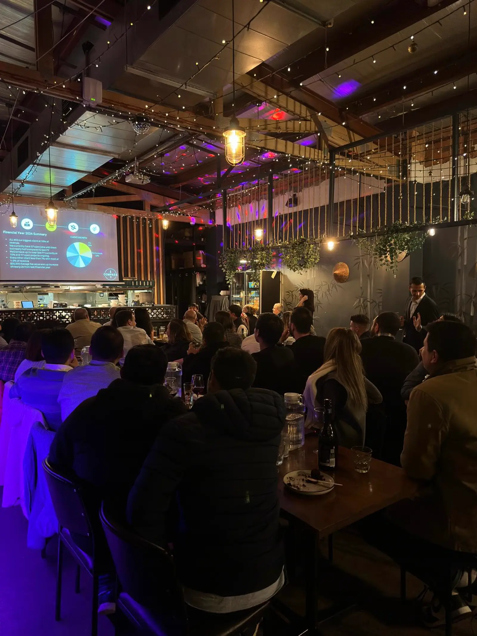 A corporate presentation on the big screen to a full dining room under fairy lights